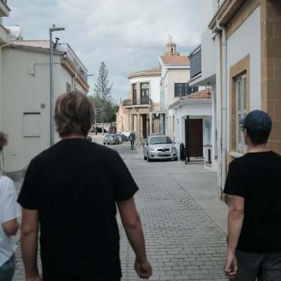 Τρεις άνθρωποι περπατούν σε πλακόστρωτο δρόμο, με κτίρια και αυτοκίνητα στο βάθος.